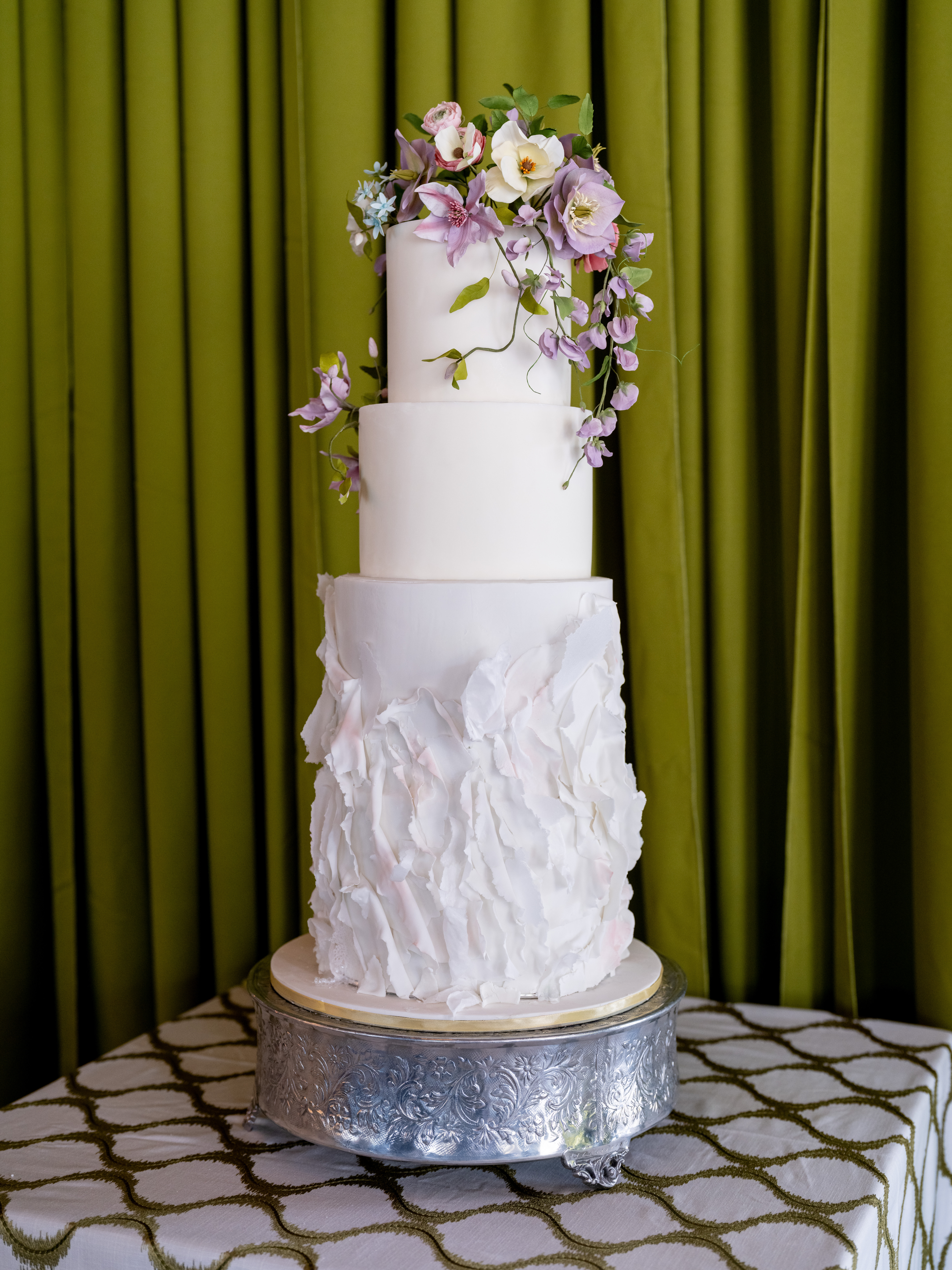 Three-tier white wedding cake with sculpted petal-textured bottom tier and cascading lavender sweet pea and ivory magnolia sugar flowers, on a silver pedestal against an olive velvet backdrop